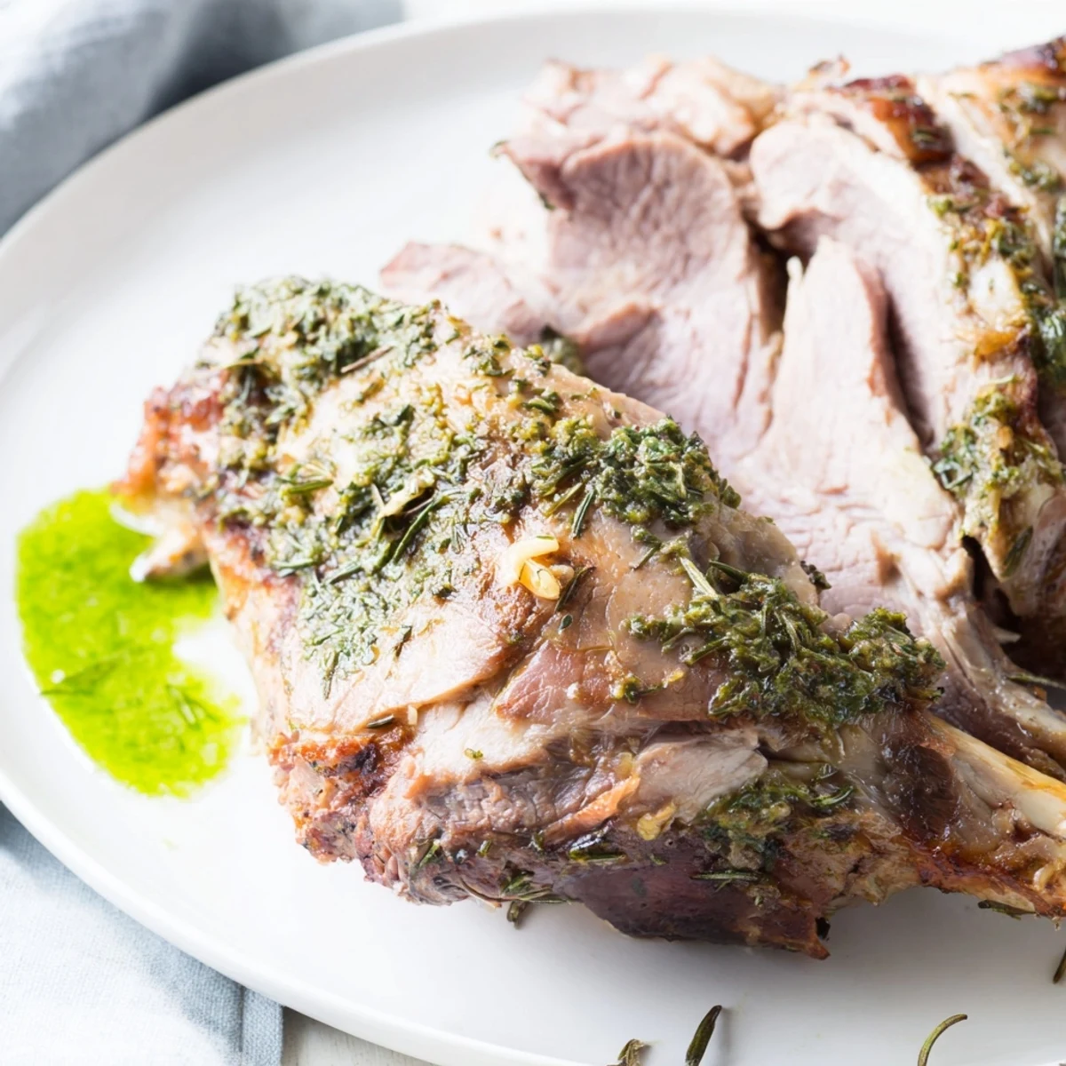 Golden-brown roasted leg of lamb with mint jelly sits sliced on a platter surrounded by rosemary sprigs and fresh mint leaves.