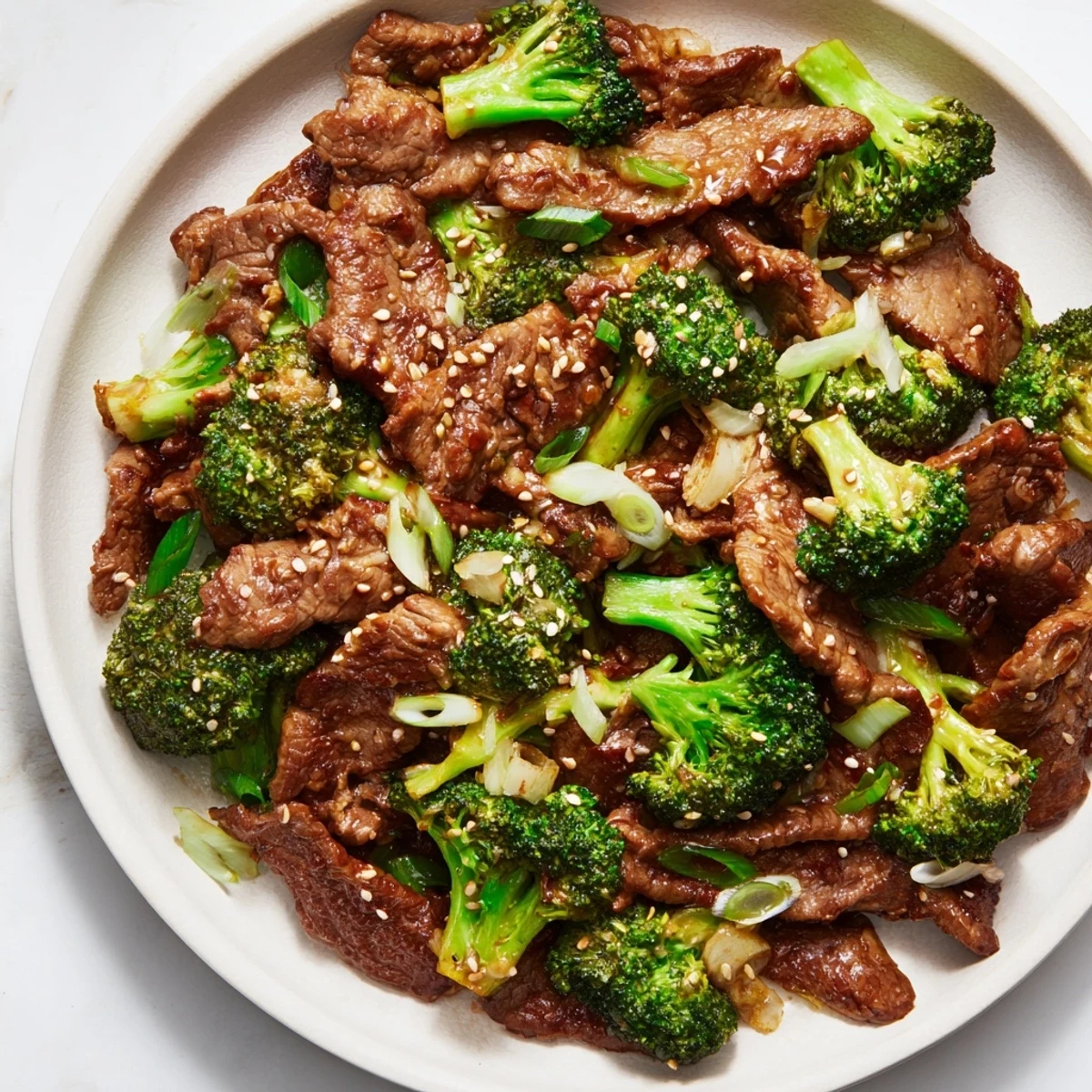 A close-up of Better Than Takeout Beef With Broccoli, featuring tender beef slices coated in glossy, savory brown sauce next to vibrant green broccoli florets, garnished with sesame seeds on a white plate.