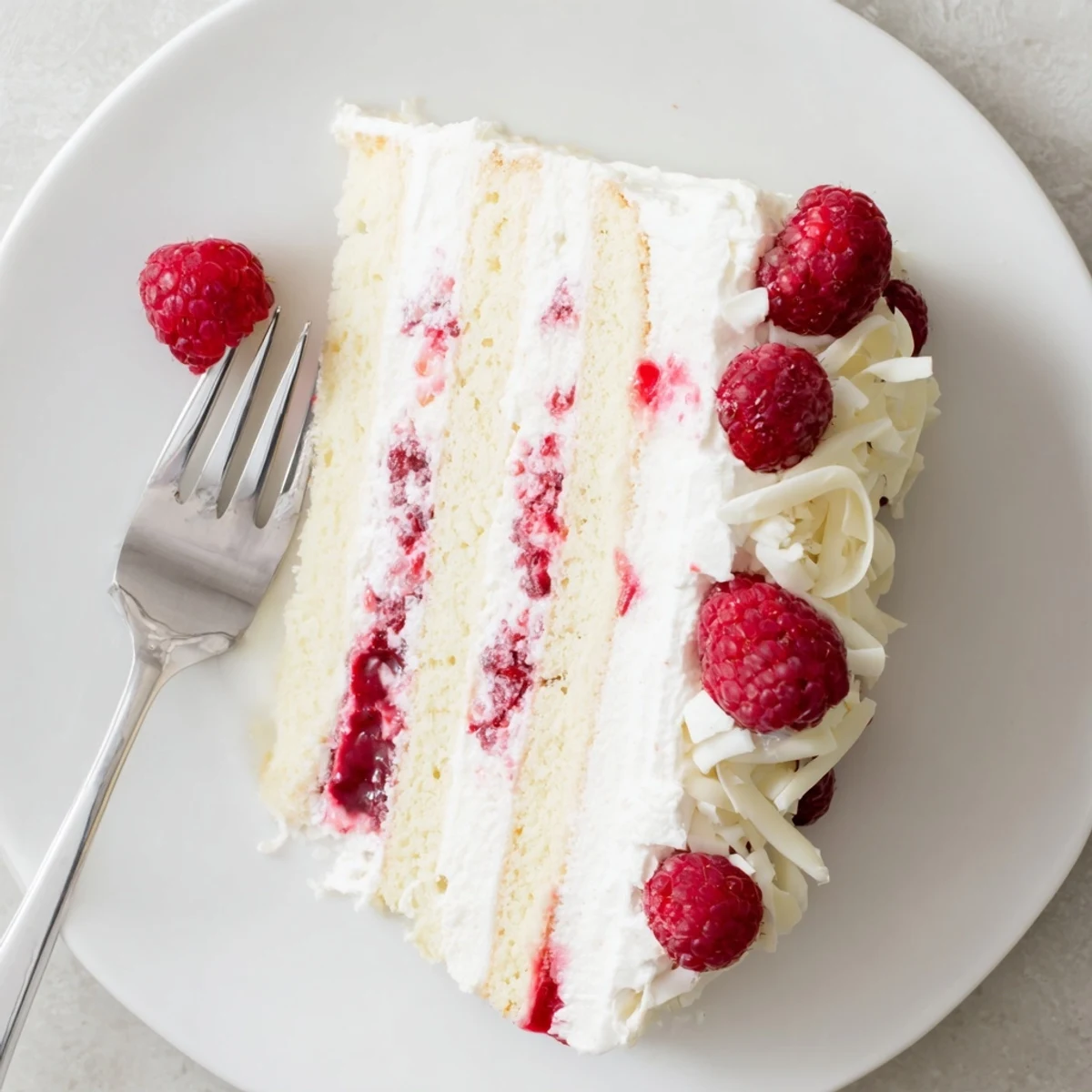 Decadent white chocolate raspberry cream cake on white plate with glossy raspberry swirls and chocolate shavings