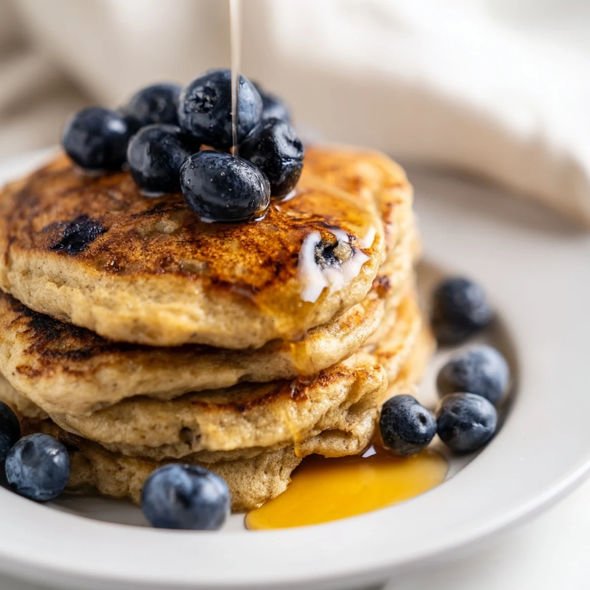 Fluffy stack of golden Greek yogurt banana pancakes topped with fresh banana slices and drizzled with warm maple syrup on a white plate.