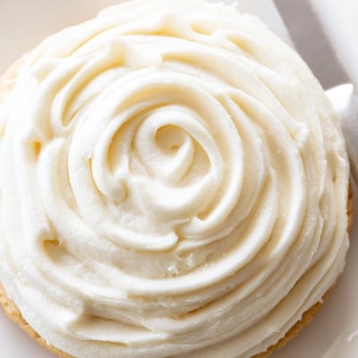 A bowl of Easy Sugar Cookie Frosting is spread on warm cookies, showing a smooth, glossy finish perfect for colorful holiday decorating.